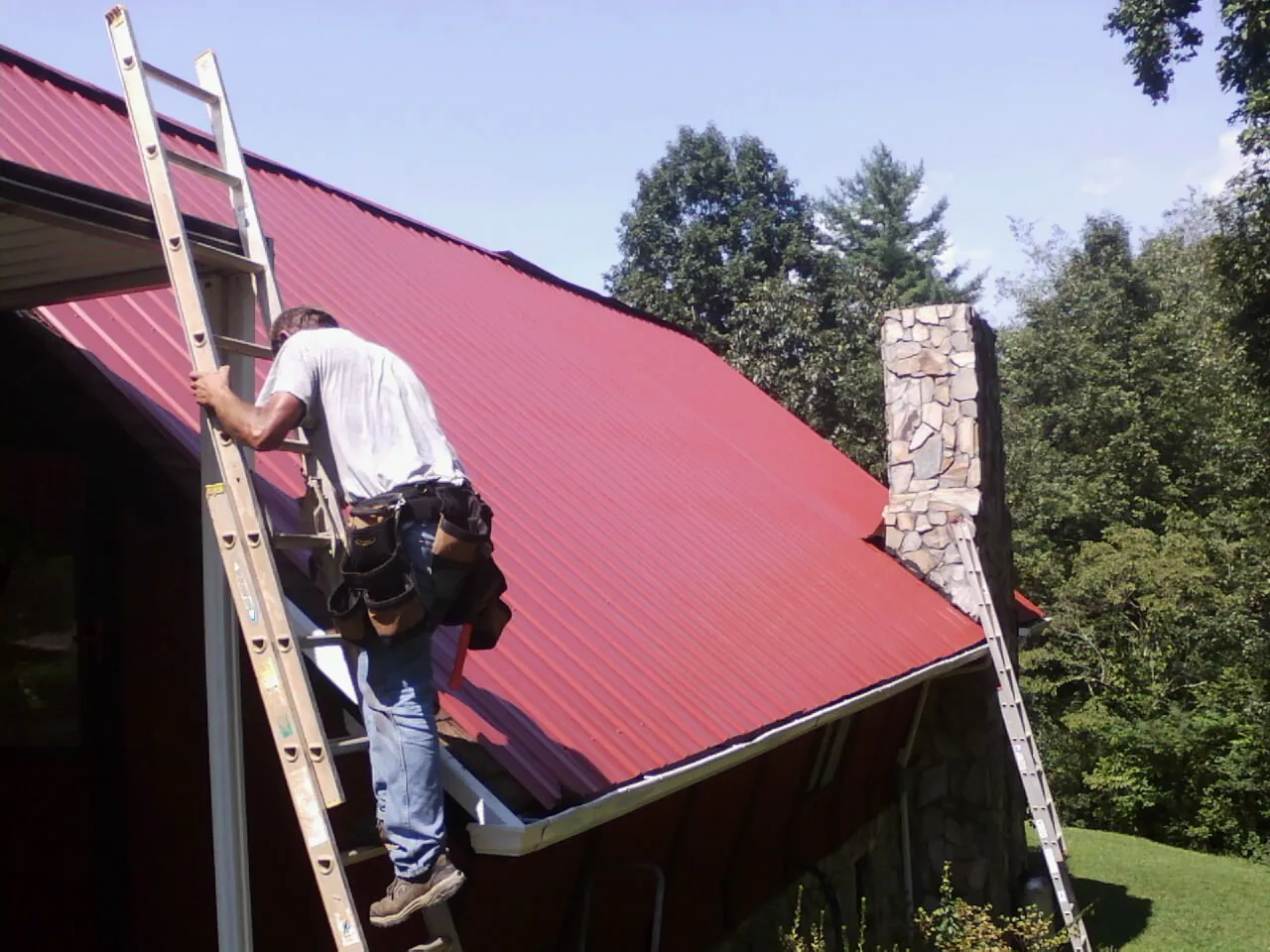 Roofer on a ladder beside a red metal roof and stone chimney — Soffit & Fascia Repair by Roofing Brothers in Miltona